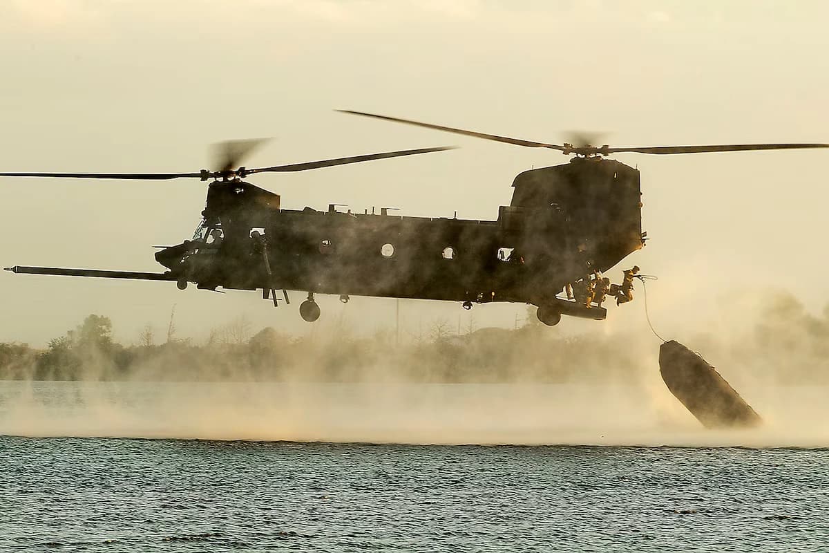 A twin-rotor military transport helicopter making a low water drop over a lake at dusk, silhouetted against a golden sky, with a crew member visible in the open cargo door.